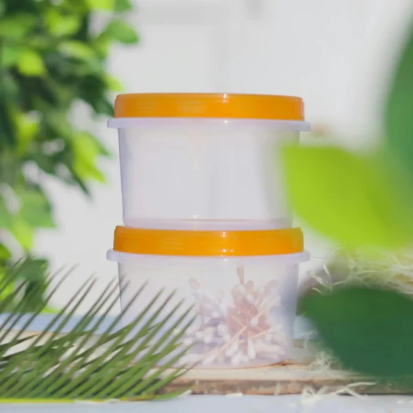 Two transparent plastic containers with orange lids stacked against a green leafy background.